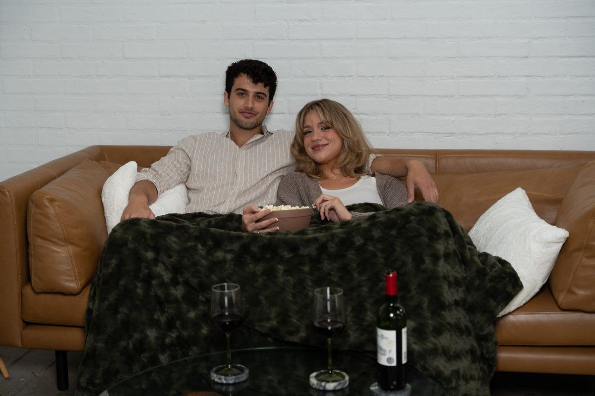 Two people on a tan leather sofa under an olive faux fur throw in the evening