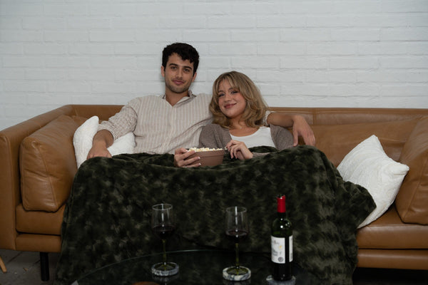 Two people on a tan leather sofa under an olive faux fur throw in the evening