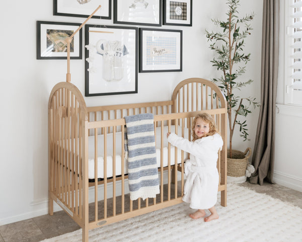 A toddler in a Keiki robe standing at a wooden crib in a bright nursery