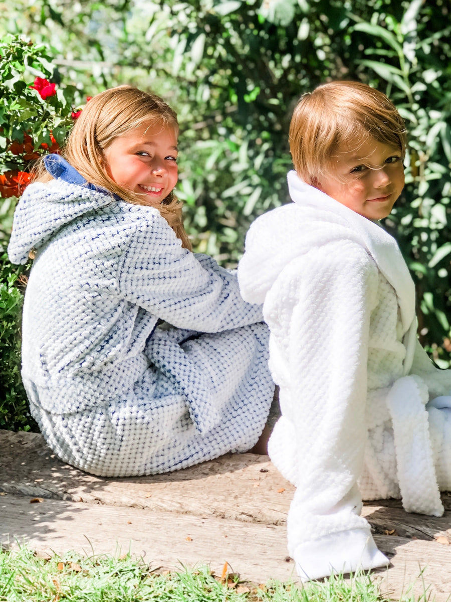 A female child wearing a white fluffy doted robe with a blue background and blue edged children's robe with a male child wearing a white children's robe who are both looking backwards and smiling.