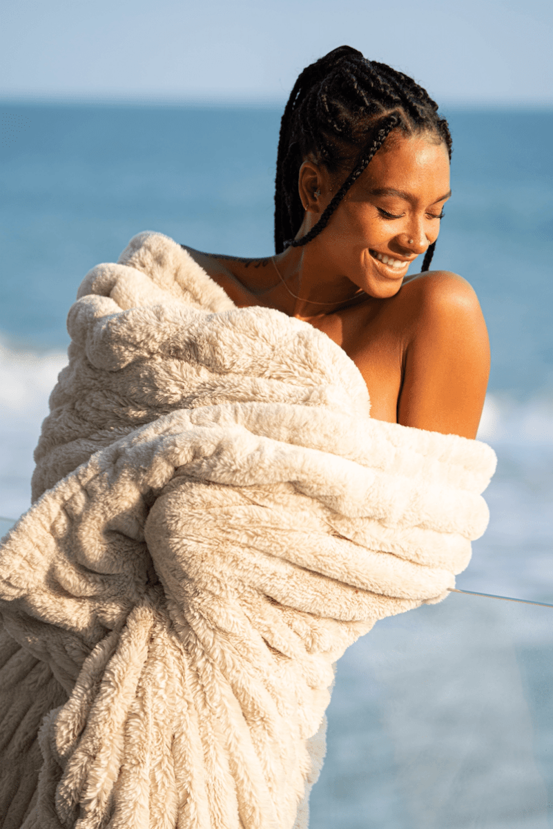 Dune Cream Cloud Throw ultra soft luxury faux fur weighted blanket wrapped around woman in luxury home by beach.